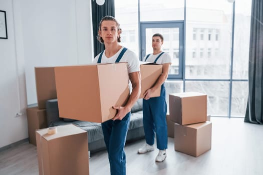 With boxes in hands. Two young movers in blue uniform working indoors in the room.