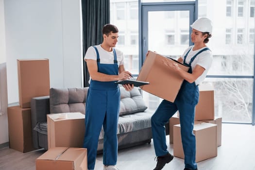 Holds notepad with document. Two young movers in blue uniform working indoors in the room.