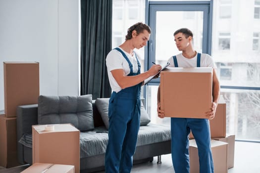 Holds notepad with document. Two young movers in blue uniform working indoors in the room.