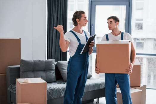 Holds notepad with document. Two young movers in blue uniform working indoors in the room.
