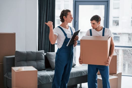 Holds notepad with document. Two young movers in blue uniform working indoors in the room.