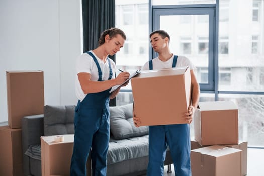 Holds notepad with document. Two young movers in blue uniform working indoors in the room.