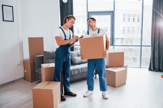Holds notepad with document. Two young movers in blue uniform working indoors in the room.
