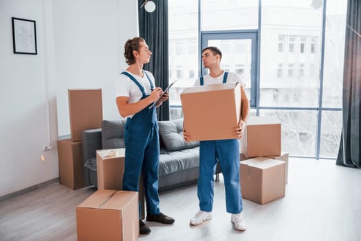Holds notepad with document. Two young movers in blue uniform working indoors in the room.