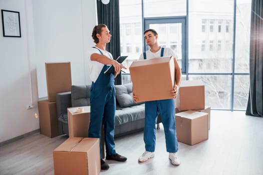 Holds notepad with document. Two young movers in blue uniform working indoors in the room.