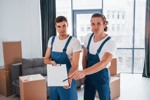 Holds notepad with document. Two young movers in blue uniform working indoors in the room.
