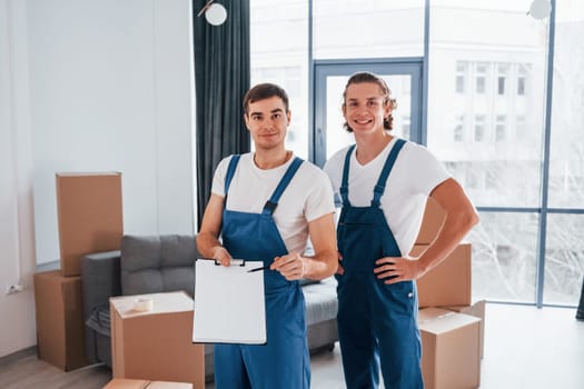 Holds notepad with document. Two young movers in blue uniform working indoors in the room.