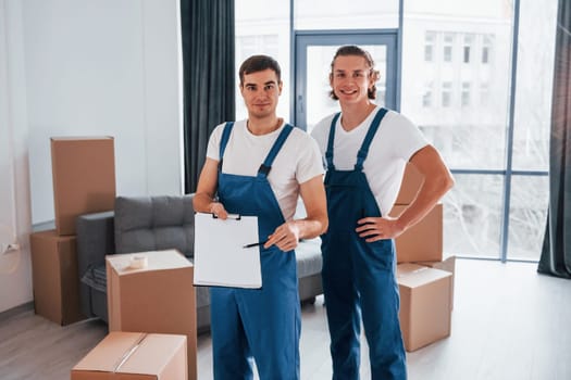 Holds notepad with document. Two young movers in blue uniform working indoors in the room.