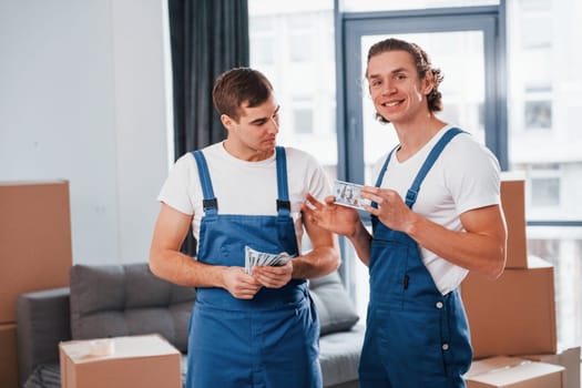 Holding money. Two young movers in blue uniform working indoors in the room.