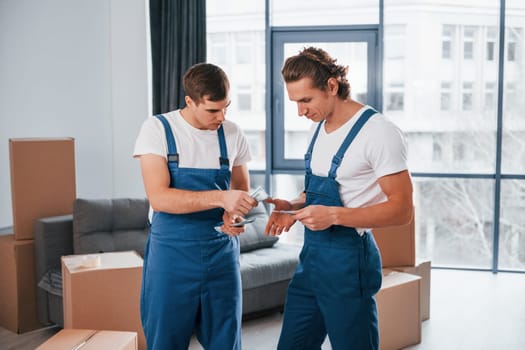 Holding money. Two young movers in blue uniform working indoors in the room.