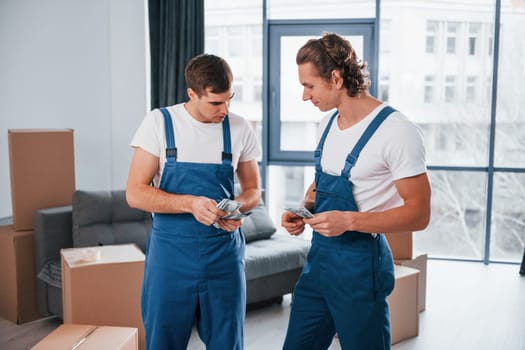 Holding money. Two young movers in blue uniform working indoors in the room.