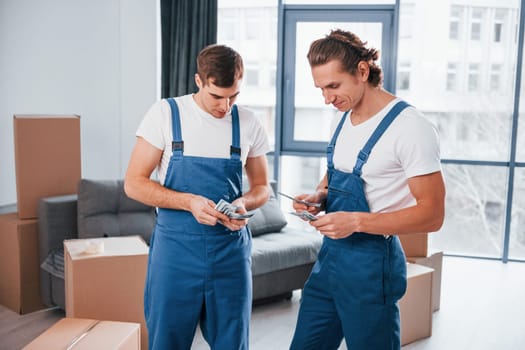 Holding money. Two young movers in blue uniform working indoors in the room.
