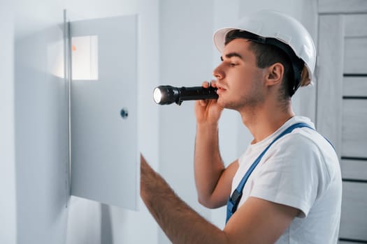 Young male electrician in white hard hat works indoors in the room.