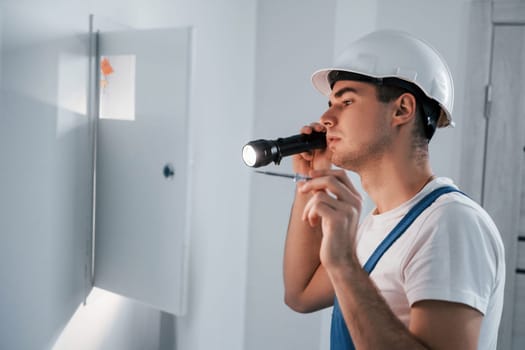 Young male electrician in white hard hat works indoors in the room.