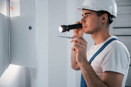 Young male electrician in white hard hat works indoors in the room.