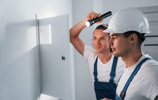 Two young male electricians works indoors together. Using flashlight.