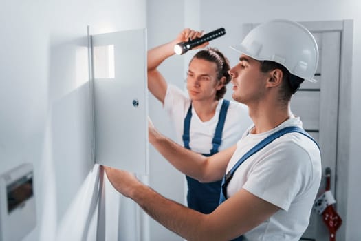 Two young male electricians works indoors together. Using flashlight.