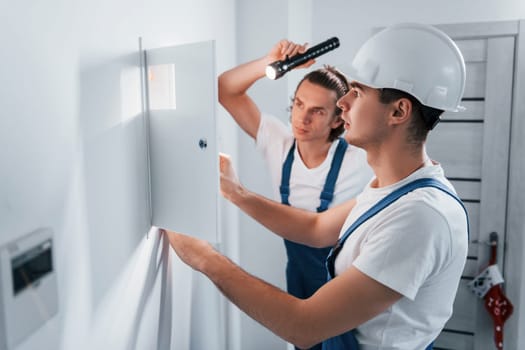 Two young male electricians works indoors together. Using flashlight.