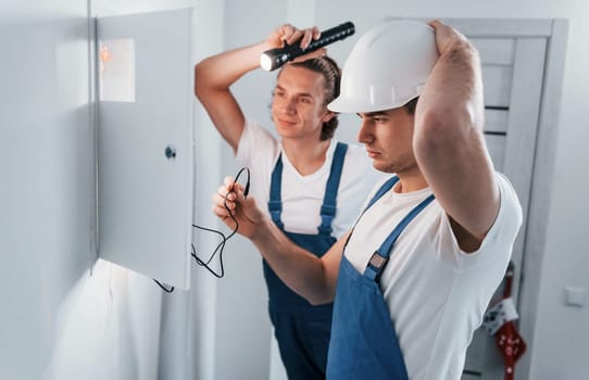Two young male electricians works indoors together. Using flashlight.