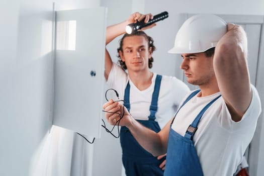 Two young male electricians works indoors together. Using flashlight.