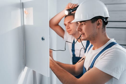 Two young male electricians works indoors together. Using flashlight.