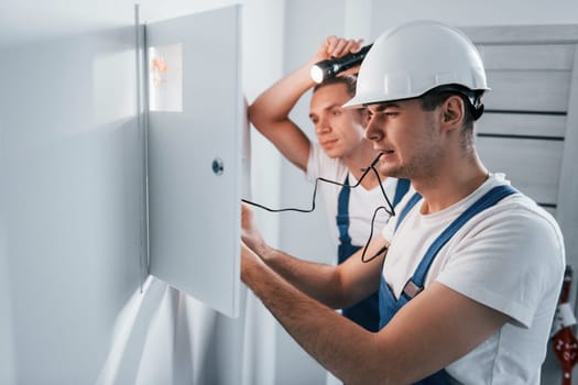Two young male electricians works indoors together. Using flashlight.