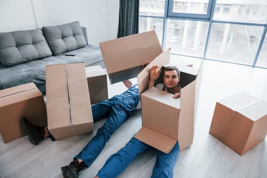 Two young male movers in blue uniform lying down on the floor with boxes.