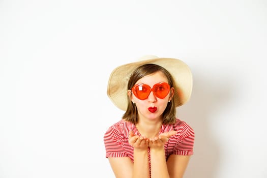 An attractive young girl with red sunglasses and summer hat sending an air kiss. Valentines day. High quality photo.