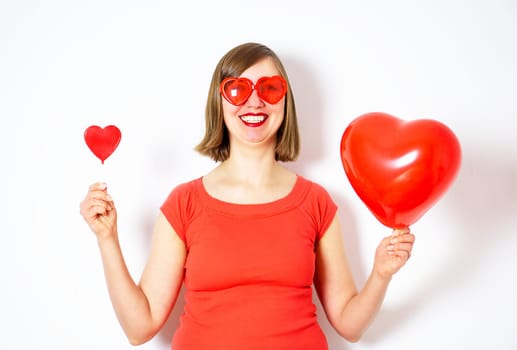Beautiful smiling woman in red sunglasses holding in her hands a red heart balloon and a red heart lollipop for Valentines Day. High quality photo