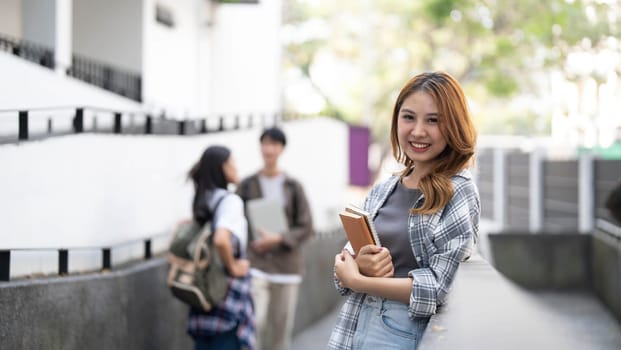 Cute asian girl student with hold a book and laptop near the campus against the background of a group of students...