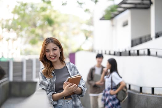 Cute asian girl student with hold a book and laptop near the campus against the background of a group of students...