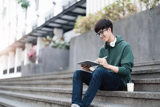 Smart Asian male college student wearing headphones, using laptop on campus outdoor stairs..