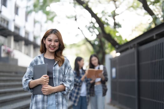 Cute asian girl student with hold a book and laptop near the campus against the background of a group of students...