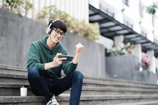 Smart Asian male college student wearing headphones, using laptop on campus outdoor stairs..