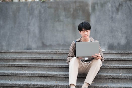 Smart Asian male college student wearing headphones, using laptop on campus outdoor stairs..