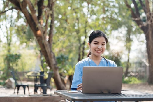 Asian female nurse working outdoor video call with patient online on laptop at desk in park...