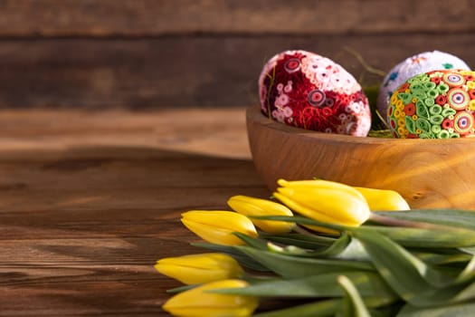 View of the Easter composition seen from the side. Patterned Easter eggs arranged on hay in a wooden bowl stand on a plank top.