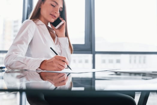 Have conversation by the phone. Young woman in white formal clothes is indoors in the modern office.