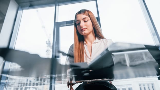 Sits by the table. Young woman in white formal clothes is indoors in the modern office.