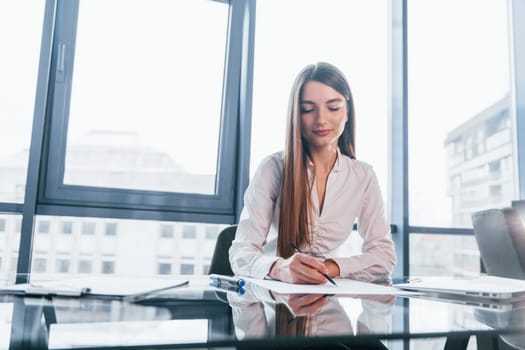 Sits by the table. Young woman in white formal clothes is indoors in the modern office.