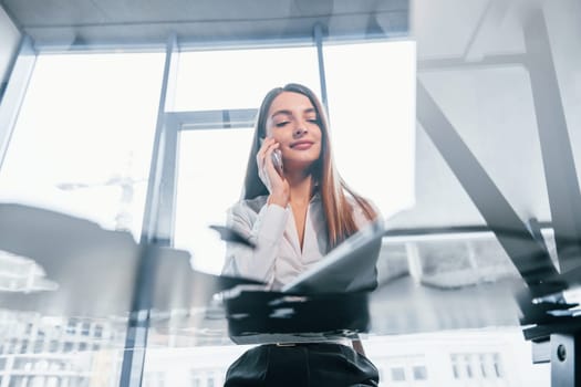 Have conversation by the phone. Young woman in white formal clothes is indoors in the modern office.