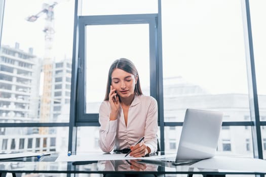 Have conversation by the phone. Young woman in white formal clothes is indoors in the modern office.