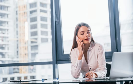 Have conversation by the phone. Young woman in white formal clothes is indoors in the modern office.