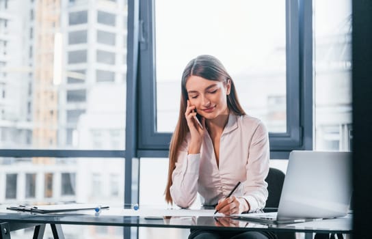 Have conversation by the phone. Young woman in white formal clothes is indoors in the modern office.