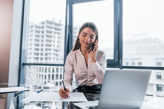 Have conversation by the phone. Young woman in white formal clothes is indoors in the modern office.