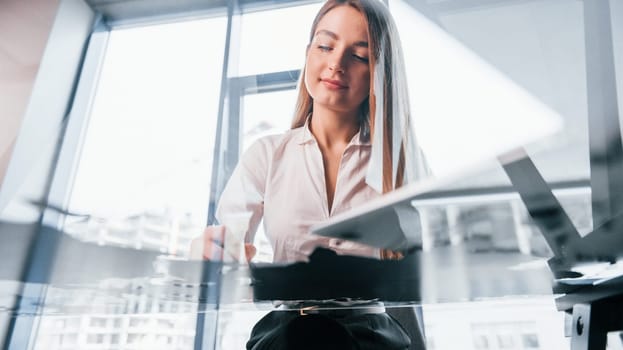 Sits by the table. Young woman in white formal clothes is indoors in the modern office.