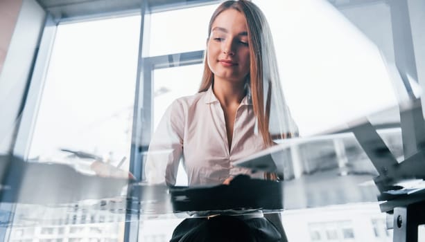 Sits by the table. Young woman in white formal clothes is indoors in the modern office.