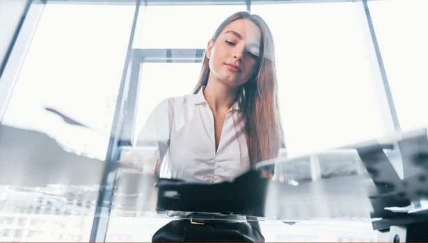 Sits by the table. Young woman in white formal clothes is indoors in the modern office.