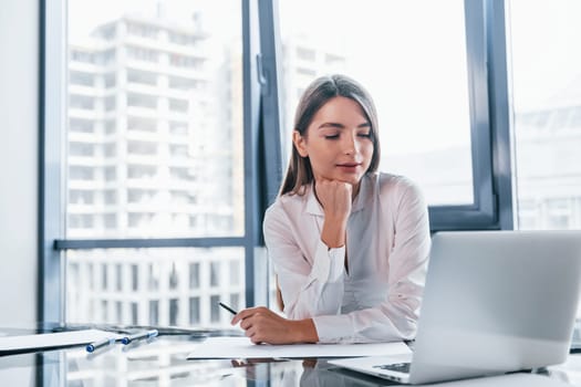 Young woman in white formal clothes is indoors in the modern office.