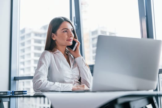 Have conversation by the phone. Young woman in white formal clothes is indoors in the modern office.